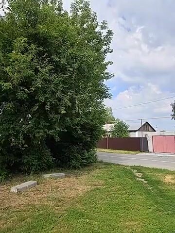 A drone shot pans right over a rural road and sidewalk, showing a large tree on the left and a pedestrian crossing with a traffic sign on the right. Houses and fences are visible in the background. The sky is cloudy and bright.