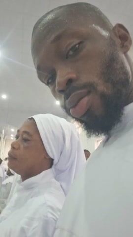 A man with dark skin and a beard is recording a video of himself in a room with several other people. All of the people are wearing white clothing. The room is brightly lit. The man is looking into the camera.