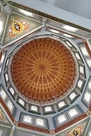 The video shows a view looking up at the dome of a building, likely a mosque, given the ornate Islamic calligraphy and geometric patterns. The dome itself is octagonal and features a rich, golden-brown color with intricate dark brown patterns radiating from a central point. These patterns appear to be stylized floral or arabesque motifs arranged in concentric circles.

The transition from the dome to the surrounding structure is marked by a series of smaller arched windows, or perhaps alcoves, set into the walls. These are separated by decorative panels that continue the intricate geometric and floral designs, as well as more calligraphy. The overall color scheme is warm, with accents of orange, yellow, and brown against a lighter, possibly gray or blue, background in some areas.

The lighting appears to be natural, filtering in from the windows, creating a sense of spaciousness and reverence. There are no people visible in the frame, and the focus remains entirely on the architectural and decorative elements of the dome and its surroundings. A thin wire or string hangs down from the center of the dome. The camera movement is a slow, steady pan or zoom, allowing for a detailed appreciation of the craftsmanship.