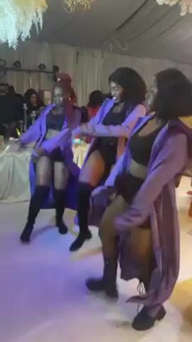 The video features three women dancing in what appears to be a party setting. They are all wearing similar outfits consisting of a black crop top, black shorts, black boots, and a long purple robe. One of the women is holding a light stick. They are moving and dancing in a coordinated manner.  The background shows party decorations, including white drapery, tables, chairs, and other partygoers. There are also lights shining on the dance floor.