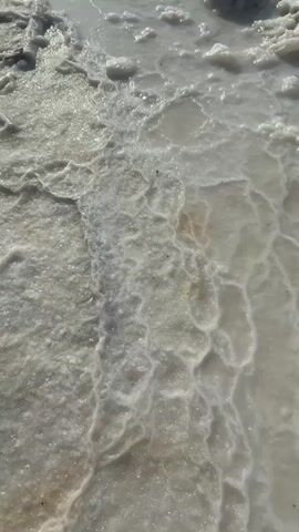 The video shows a close-up of salt formations on the shore of a body of water. The water is a clear, greenish hue, and the seabed is visible, with rocks present. In the distance, there is a faint outline of land and a structure in the water. The sky is a clear blue with a few wispy clouds. The overall scene suggests a natural environment, likely the Dead Sea, given the prevalence of salt deposits and the water's unique color. There are no people or animals visible in the video. The camera is positioned low, near the water's edge, and pans across the salt formations and then out towards the open water.