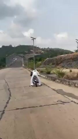 A person wearing a white outfit and a black letter on their back is seen on a motorcycle at the start of a paved road. The motorcycle then starts forward and moves quickly down the road, away from the camera. The road leads to a bridge with a metallic frame and wire mesh on either side. The surrounding landscape includes green hills and visible signs of construction or excavation. The sky is overcast. The person on the motorcycle is the only individual visible.