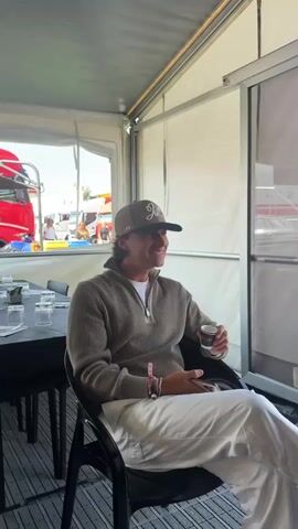 The video shows a man sitting in a tent with white fabric walls and a clear plastic wall section. He is wearing a beige quarter-zip sweater over a white t-shirt, white pants, and a beige baseball cap with a dark brim and a logo on the front. He holds a small cup in his right hand and a smartphone in his left hand. He looks to his right and smiles, then turns his head slightly and appears to be speaking or reacting to something off-camera. In the background, through the clear plastic wall, a red truck and other vehicles are visible, suggesting an outdoor event or venue. The table in front of him has glasses and a black tablecloth.