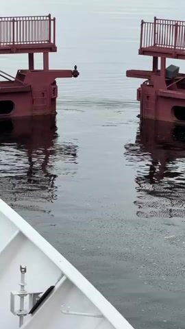 The video begins with a close-up of water with small bubbles, showing the reflection of a red structure with railings. The camera then pulls back to reveal a body of water with two red structures, possibly part of a lock or bridge, spanning across it, with a gap in the middle. The bow of a white boat is visible in the foreground, with a coiled black rope and some equipment. The water is calm, with ripples indicating movement. The sky is overcast, and a distant shoreline with trees and buildings can be seen. The camera continues to pull back, showing more of the surrounding water and landscape. The overall scene depicts a boat approaching or departing from a waterway controlled by a lock or bridge mechanism.