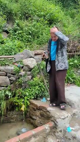 A woman stands in front of a natural spring in a lush, green environment. She is wearing a blue t-shirt, a patterned jacket, and brown pants. She is holding a bottle in her right hand and appears to be wiping her eyes with her left hand. The spring water flows into a stone basin, and some debris, including plastic bags, is visible near the water's edge. The background is filled with dense vegetation.