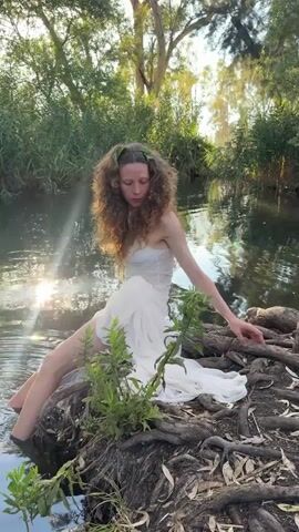 A person with curly brown hair adorned with green leaves is sitting on exposed tree roots at the edge of a body of water. They are wearing a white, strapless dress with a sheer bodice and a flowing skirt. Their legs are partially submerged in the water, and their right arm is extended towards the water's surface. Green vegetation grows around them, and the background consists of trees and sunlight reflecting on the water. The person is looking directly at the camera.