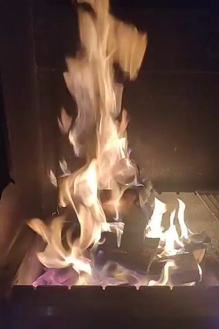 The video shows a close-up shot of a fire burning in a barbecue pit. The flames are a mix of yellow and orange, with some areas appearing white hot. The fire is consuming several pieces of wood, which are glowing red from the heat. Sparks are seen flying from the fire, and the background is dark, indicating that it is nighttime. The video is shot from a fixed perspective and appears to be handheld, with some slight camera shake.