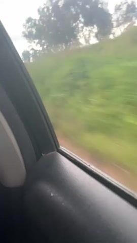The video shows a view from inside a car, looking out the window. The car appears to be moving, as indicated by the motion blur of the scenery outside. The scenery consists of a dirt road, green grass and vegetation, trees, and a cloudy sky. In the distance, there is a small structure that looks like a shelter or a shack. The interior of the car, including the door panel, is partially visible in the foreground. The audio consists of a conversation in a language that appears to be Swahili, where the speakers express dislike for something and discuss reporting an issue.