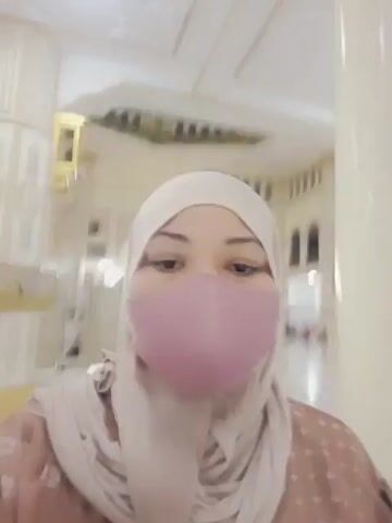 The video shows a woman wearing a light pink hijab and a matching face mask, holding a camera to take a selfie. She is in a large, open indoor space with white tiled floors and tall, ornate columns. The lighting is bright, suggesting it's daytime or well-lit indoors. In the background, other people can be seen in the distance, some appearing to be sitting or lying on the floor, and architectural details suggest a place of worship or a grand public building. The overall atmosphere appears calm and reverent. There is no visible text in the video.