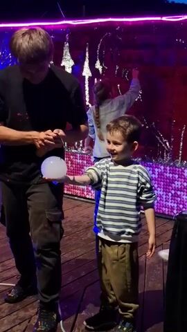 On a wood plank floor, a man in black clothing uses a small torch to light a spherical lamp held by a young boy in a striped shirt and brown pants. A woman in a red dress smiles nearby. Behind them is a wall with reflective panels and a decorative backdrop with purple and blue lighting. Another child stands by the wall. The lighting in the scene is dim. The boy is smiling, and the woman looks pleased.