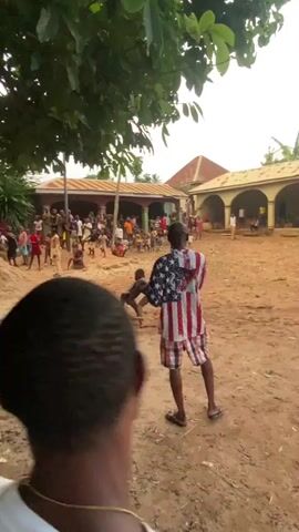 The video captures an outdoor scene in what appears to be a village or community courtyard in Unwana, Nigeria. At the center of a large, dry dirt area, two young boys are engaged in a wrestling match, crouched low and facing each other. A crowd of numerous people, largely composed of men and boys, surrounds the wrestling pair, actively observing the event.

In the mid-ground, with his back to the camera, a man wearing a distinctive American flag-patterned short-sleeved hoodie and matching shorts is holding a smartphone, seemingly recording the wrestling. Another person is visible in the immediate foreground, wearing a baseball cap with a "Gucci" logo clearly visible on its side.

The background features several single-story buildings characterized by red roofs and arched doorways, consistent with local architecture. Green foliage from overhead trees provides some shade over the area. The ground is dry and dusty, with a few pieces of litter scattered. The overall impression is that of a community gathering, with spectators engrossed in the informal wrestling match taking place under daytime, possibly late afternoon, lighting conditions.