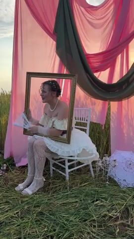 The video features a person sitting on a white chair outdoors, holding a golden frame in front of their face. They are wearing a white dress with ruffles, white patterned stockings, and white sandals. The person also wears gloves and a pearl necklace. A fan is visible in their hands. Behind the person, pink and green fabric drapes are set up, creating a backdrop. The scene is set in a grassy field, and the sun appears to be setting, casting a warm glow. A white umbrella with lace detailing is visible on the right side of the frame. The person looks in various directions and smiles at the camera at one point.