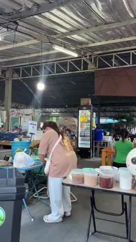 The video pans across an outdoor covered market or food stall area. In the foreground, to the left, a woman with long dark hair, wearing a beige t-shirt and white pants, is seen from behind, preparing something at a blue table. She has a pink bag slung over her shoulder. To her right, a folding table displays several small containers of food items and bowls. In the mid-ground, a refrigerator unit stocked with drinks is visible. Several people are seated at tables, eating and socializing. A woman in a green shirt is sitting at one table. Further back, a man is seen eating at a wooden table. A Thai flag is prominently displayed, hanging from a pole with a blue light fixture attached. Motorcycles are parked in the foreground and mid-ground. The environment appears to be a bustling open-air eatery.