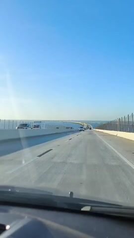 The video is a first-person perspective shot from inside a vehicle driving on a bridge over a body of water. The sky is clear and blue. The bridge has multiple lanes and is lined with railings and fences. There are other vehicles on the bridge, including cars and a truck. The water is calm and stretches to the horizon. The sun is shining, casting shadows on the bridge.