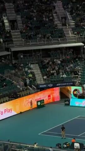 The video shows an overhead shot of a tennis match in a stadium with many spectators. A tennis court with blue lines is visible in the foreground. Two players in orange outfits are seen on the court. Numerous people are seated in the stands, which are arranged in tiers. Electronic billboards displaying "Miami Open" and "Where Tennis Comes To Play" are located along the perimeter of the court. Several individuals, possibly referees or staff, are seen standing around the court. Cameras and technical equipment are also present at the bottom of the frame.