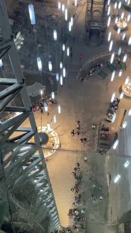 The video shows the interior of a large, cavernous space, possibly a salt mine, lit with many hanging cylindrical lights. The walls of the space are a rough, gray texture, and the ceiling is high and appears to have a layered or striated pattern. Several platforms and walkways are visible within the space, with people walking around and gathering in small groups. There are structures like small tables and a circular stage-like area. The video pans upwards to show the full vertical extent of the space. A large metal structure can be seen on the left. The overall impression is of a subterranean, illuminated space repurposed for public use.