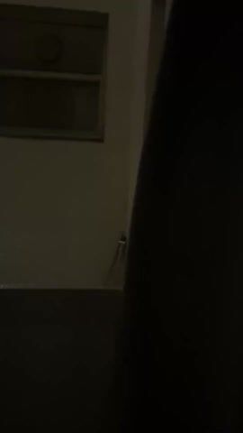 The video depicts a dimly lit room with a window partially visible. The window is square and has a round vent near the top. The surrounding walls are a light color, possibly beige or off-white. A dark object, potentially a curtain or wall, is prominent on the right side of the frame.  The camera pans slightly upward, focusing on the window. The overall scene is dark and the details are not clearly discernible.
