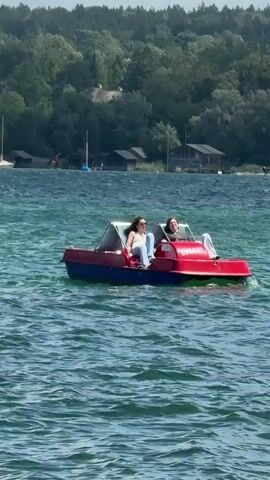 Two young women are in a red and blue pedal boat on a lake. The woman in the front is wearing a white top and blue jeans, and the woman in the back is wearing a grey top and jeans. They are both looking to the left. The boat is red with a blue hull and a clear canopy. The water is choppy, and there are trees and buildings on the shore in the background. The sky is blue with some clouds. The video is a still image with no discernible motion.