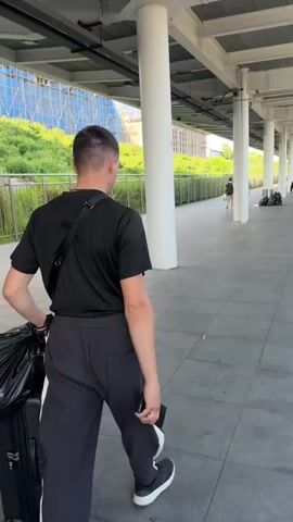 The video shows a person walking with luggage at a train station platform. The first shot shows the back of a man in a black t-shirt and black pants with white stripes walking away from the camera, pulling a black suitcase. He has a black bag slung across his shoulder. The second shot pans to the right, showing a train at the platform and other people with luggage waiting. The third shot is a close-up of the tiled floor and the yellow tactile paving at the edge of the platform. The fourth and fifth shots are from a first-person perspective, showing someone holding a white suitcase and a brown handbag with red trim, also on the platform. The environment appears to be an outdoor train station with a covered platform, visible pillars, and some greenery and buildings in the background.