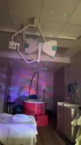 A woman in a red shirt is dancing in a birthing room. The room has a bed, a birthing tub, and medical equipment hanging from the ceiling. There are colorful lights projecting onto the walls and ceiling, creating a festive atmosphere. The woman's back is to the camera.