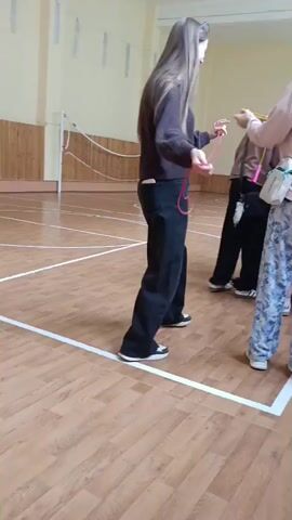 The video shows a group of young people in what appears to be a gymnasium. The floor is polished wood with white lines marking boundaries. The walls are a light color, and there are windows with a textured glass or screen visible. The lighting suggests it is daytime.

Several individuals, mostly teenage girls, are interacting with sports equipment. In the foreground, a girl with long, dark hair tied back is holding a red jump rope, appearing to be in motion with it. Another girl, wearing a light pink sweatshirt and patterned pants, stands facing away from the camera, holding a white bag with a green strap. To her left, another girl in a dark hoodie and jeans is holding what looks like a tablet or book. Further back, another girl in a dark outfit is holding a jump rope. The scene suggests a sports class or a recreational activity where participants are being given or are using jump ropes, tennis balls, and possibly other sports equipment. The overall mood seems relaxed and casual, with some individuals engaged in activity and others observing or preparing. No text is visible in the video. The context provided places this scene in Penza, Russia.