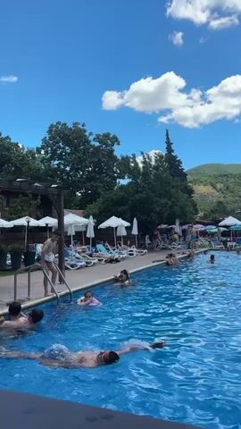 A long shot captures a crowded outdoor swimming pool on a sunny day. In the foreground, a man swims towards the left of the frame. To his right, the water is filled with people swimming and wading. Along the edge of the pool, lounge chairs are occupied by sunbathers, some under umbrellas. A pool ladder is visible on the far left. In the background, lush green hills rise under a bright blue sky with scattered white clouds. Trees and some buildings with red tile roofs are interspersed with the hills.