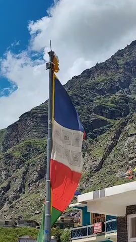A prayer flag, with blue, white, red, and green panels, flutters from a metal pole against a backdrop of a steep, rocky mountainside under a blue sky with white clouds. Below and to the right of the flag, a building with a light blue and white facade is visible. Additional prayer flags can be seen further in the distance, strung along a ridge. The scene appears to be in a mountainous, possibly Himalayan, region.