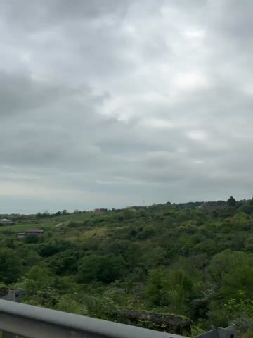 The video shows a landscape on a cloudy day. There are green, tree-covered hills with scattered houses and buildings in the distance. The sky is overcast with grey clouds. A roadside guardrail is visible in the foreground.