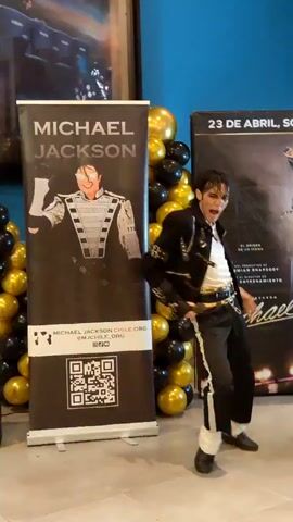 A person dressed as Michael Jackson is performing a dance routine. They are wearing a black sequined jacket, white shirt, black pants with a white stripe, and white socks with black shoes. The performer is holding a microphone in their right hand and their left arm is extended to the side. They are in front of a banner that says "MICHAEL JACKSON" and a wall decorated with black and gold balloons. Another poster is visible to the right of the performer.