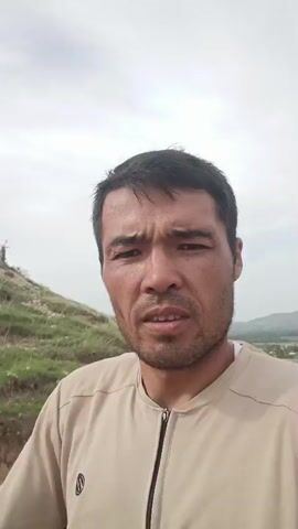 A man is riding a horse on a dirt road. He is looking at the camera and talking. The horse is brown and has a saddle. The man is wearing a light brown shirt and blue jeans. The road is surrounded by hills and grass. The sky is cloudy.