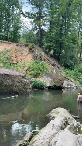 A person jumps from a dirt embankment into a body of water surrounded by trees and rocks. The person is wearing shorts and a dark top, and appears to be mid-air before entering the water. Another person, also wearing shorts, stands in the water observing the jump. The water is murky and reflects the surrounding greenery. Large rocks are present in the foreground and along the water's edge. The embankment has exposed roots and vegetation.