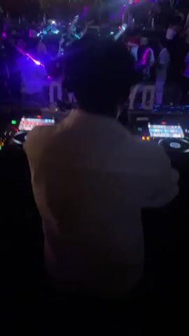 The video shows a DJ booth in a dimly lit nightclub. The DJ, seen from behind, is wearing a white shirt and has dark, curly hair. They are operating professional DJ equipment, including two turntables and a mixer, with various lights and displays. In the background, a crowd of people are dancing and enjoying the music, illuminated by colorful club lights, including purple and blue. A prominent sign behind the DJ reads "DYNAMO" in glowing letters. The overall atmosphere is energetic and typical of a nightlife venue.