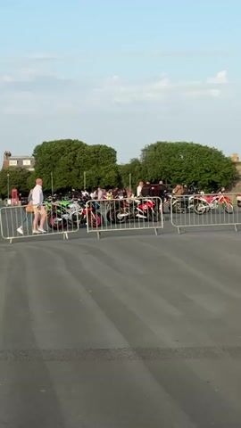The video shows a partially-fenced area with several motorcycles parked inside, along with a number of people standing around. A man and woman walk past the fenced area. The camera pans to the right to show a row of flags. Beyond the flags is a building and what appears to be an ice cream van. The sky is clear, and the sun appears to be setting.
