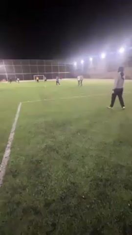 The video shows a group of people playing soccer on a green field at night. The field is lit by several bright lights. A man in a green and white striped shirt and gray pants is preparing to kick a soccer ball. Another man wearing a yellow jacket is standing behind him. There is a soccer goal visible in the background. Several other people are scattered around the field.
