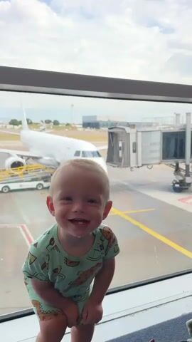 The video begins with a close-up shot of a smiling toddler looking up at the camera. The toddler has blond hair and is wearing a light green t-shirt with a pizza and dinosaur pattern. The background is an airport window with an airplane visible outside. The toddler then turns to look out the window, leaning forward to get a closer look at the airplane. They appear to be pressing their face against the glass, and a small amount of liquid, possibly drool, is visible on the window near their mouth. The toddler then turns their back to the camera and raises one hand as if to wave at the airplane. The scene is shot from a slightly low angle, emphasizing the toddler's perspective.