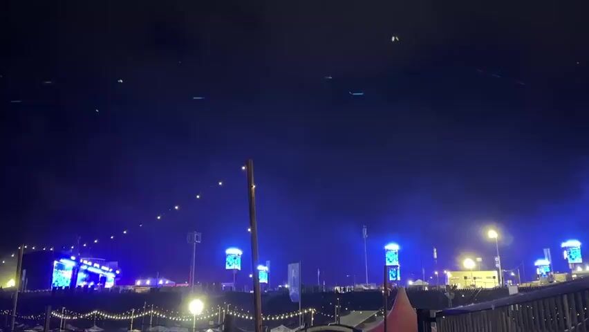 The camera captures a dark night scene at an outdoor event, possibly a music festival, before quickly shifting downwards into a dark, blurry view. The initial shot shows a wide, open area illuminated by various lights. In the distance, a large stage with bright blue and white lights is visible, positioned on what appears to be a raised platform or hill. Several tall structures, possibly light towers or screens, with glowing blue displays are scattered across the midground. String lights are draped across the foreground, creating a festive atmosphere. A "TOILETTES" sign is faintly visible on a portable toilet in the bottom left, indicating facilities. A red-orange tent is also visible on the right. The sky above is dark, with some faint, moving green lights or possibly lasers. The ground in the foreground is uneven, suggesting a natural or unpaved surface. The camera then abruptly moves downwards, resulting in a dark, blurry, and undecipherable view for the remainder of the clip.