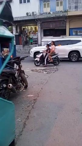Two people are on a motorcycle on a paved street. The person in the back is wearing an orange shirt and green shorts. The person in the front is wearing a black shirt and white pants. There are buildings and other vehicles in the background. The motorcycle appears to be moving. A pothole is visible in the road.