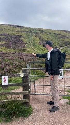 A man wearing a black jacket, white t-shirt, tan pants, a black baseball cap, and glasses stands in front of a metal gate. He is holding a black walking stick and looking towards the gate. Behind him is a hill covered in purple heather and a path leading up the hill. There are two signs on the wooden fence to the left of the gate, one of which is a no dogs allowed sign. The man is also wearing a black fanny pack.
