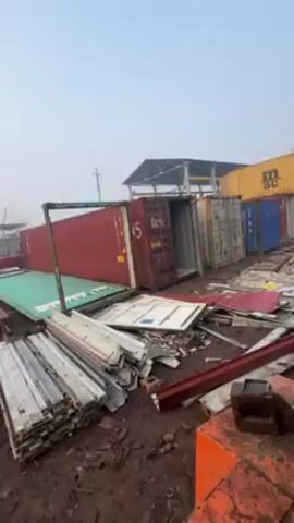 The video shows a view from inside the cab of a forklift. The forklift is parked in a storage yard with shipping containers and debris scattered on the ground. The inside of the cab is dusty and worn. The windows are dirty and fogged. The steering wheel and control levers are visible. The forklift is stationary. The environment is industrial and appears to be outdoors, judging by the overcast sky.