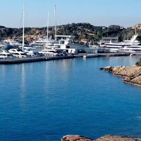 The scene depicts a marina filled with yachts and boats. The water is a deep blue and reflects the clear sky above. The boats are mostly white, and a mix of motor yachts and sailing vessels are visible. Some of the boats are docked along a pier, while others are anchored in the marina. In the background, there's a land mass with rocky terrain and sparse vegetation. There is a clear structure on the pier with what appears to be a control tower on top.
