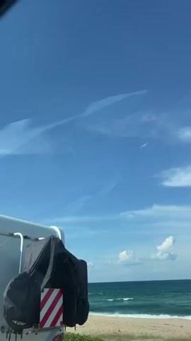 This video shows a white caravan parked on a beach next to the sea. The sky is blue with some clouds. A kite is flying in the sky above the sea. There are also some plants and bushes on the beach.