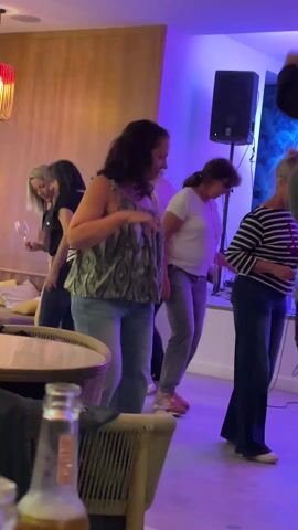 The video shows several women of various ages dancing at an indoor party with a band playing live music. The women are all smiling and appear to be having a good time, and are dancing in place. The interior is casually decorated, with a mix of modern and rustic elements. There are tables and chairs scattered around. A beer bottle sits on a nearby table. The lighting includes colored lights adding to the festive atmosphere. A speaker and microphone stand are visible near the wall.