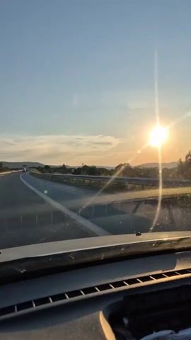 The video shows a view from inside a car traveling on a highway at sunset. The sun is visible in the upper right corner, casting a bright glare. The road stretches ahead, with some other cars in the distance. The sky is clear with a few wispy clouds. The interior of the car, including the dashboard and windshield, is visible in the foreground.