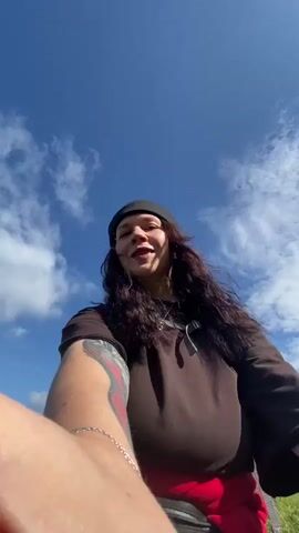 The video begins with a low-angle close-up of a person wearing a black beanie and a brown shirt, with a red garment visible around their waist. They have long, dark, curly hair and a tattoo on their arm. The person is smiling and looking at the camera, with a wide blue sky and white clouds in the background. The camera then pans out to reveal a lush green landscape with rolling hills and a distant village. The person remains in the foreground, occasionally touching their face or adjusting their position. Two other individuals can be seen in the background, also appearing to be hiking or walking. The scene suggests an outdoor activity in a natural setting, likely a hike or a visit to a scenic viewpoint.