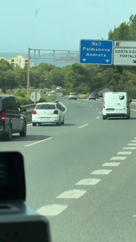 The video shows a view from inside a vehicle traveling on a highway. The highway has multiple lanes and is surrounded by green trees and some buildings in the distance. There are several cars and vans on the road. A blue road sign indicates directions for "Palmanova Andratx" and "Ma-1". The weather appears to be clear and sunny.