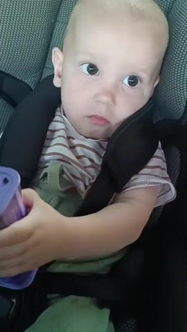 A young child is sitting in a car seat. They are holding a small, purple electronic device. The child looks at the device and then turns their head to the side. The child's hands are visible as they hold the device. The background of the video is out of focus but appears to be the interior of a vehicle.