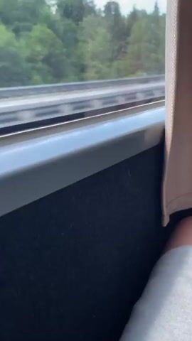 The video shows a first-person perspective of someone riding on a bus or train. The person is sitting by the window and wearing gray shorts. Outside the window, there is a highway with cars passing by, and a forest in the background. The sky is blue with white clouds. Other passengers can be seen in the seats ahead. The interior of the bus or train is visible, including the seats, curtains, and storage pockets.