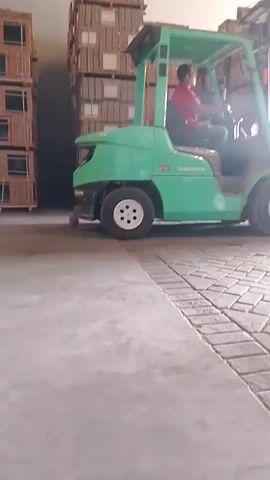 A person wearing a red shirt and a mask is operating a green forklift in what appears to be a warehouse. The forklift has the word "GRENADIER" visible on its side. Tall stacks of boxes or pallets are visible in the background. Another person, wearing a dark shirt and shorts, is standing near the forklift's forks. The forklift's forks are raised and appear to be engaging with a pallet of goods. The forklift is on a paved surface with a distinctive brick-like pattern. The lighting suggests an indoor environment with artificial light sources. The video is a short, continuous clip showing the forklift moving and interacting with the pallet.