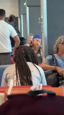 The video shows a waiting area with several people sitting and standing. In the foreground, a person with long braids and headphones sits facing away from the camera. Behind them, a man wearing a blue beanie and a fake beard looks directly at the camera with an expression of amusement. A young girl stands behind the man with the beard and looks toward the camera, mouth open in what appears to be a gasp. To the right of the man with the beard, another person with curly gray hair sits and talks with hand gestures. Several other people are seen in the background, some walking and others standing. The area appears to be an airport waiting area, indicated by the seating arrangement and the presence of other travelers. The lighting is bright and appears to be from overhead fluorescent lights. The scene is observed from a seated position.