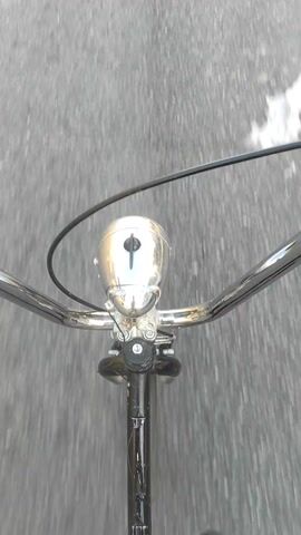 The video opens with a close-up, top-down view of a bicycle's chrome headlight and handlebars, moving over a blurry grey surface, likely a road.

From 0:02, the perspective shifts to a forward-facing view from the moving bicycle, revealing a wide, paved street in what appears to be an urban or suburban area. The street is lined on both sides by various buildings, some resembling residences and others commercial establishments. Numerous utility poles with overhead wires are prominent along the street. Several cars are parked along the left side of the road.

Long shadows, including a distinct shadow of a bicycle and its rider, are cast across the asphalt from the right, suggesting either early morning or late afternoon. The sky is clear and blue with scattered white clouds, indicating sunny weather.

Visible text on signs along the left side of the street includes "FERTILIZANTES" and a brand name (possibly "MAMINA") on a white bag, a partially obscured red and white sign (possibly "ALTO" or "ATCO"), a sign for "MADERERA" (Lumberyard), and a "CORRALON" (Building materials supplier) sign featuring yellow text and a red logo. The bicycle continues its forward movement along the street.
